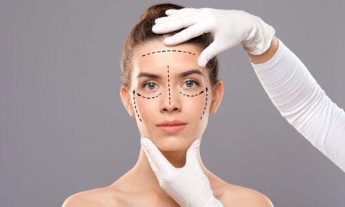 Concept of plastic surgery. Young woman getting treatment at cosmetology clinic, changing her appearance. Plastic surgeon making marks on beautiful lady face, grey studio background, copy space